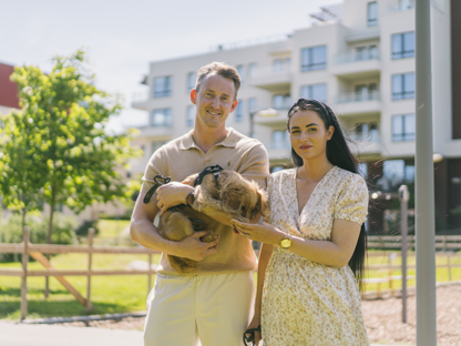 Dame og mann som står med en hund i hendene utenfor et boligområde i byen. Sol og sommer. 