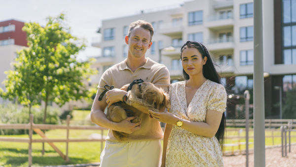 Dame og mann som står med en hund i hendene utenfor et boligområde i byen. Sol og sommer. 