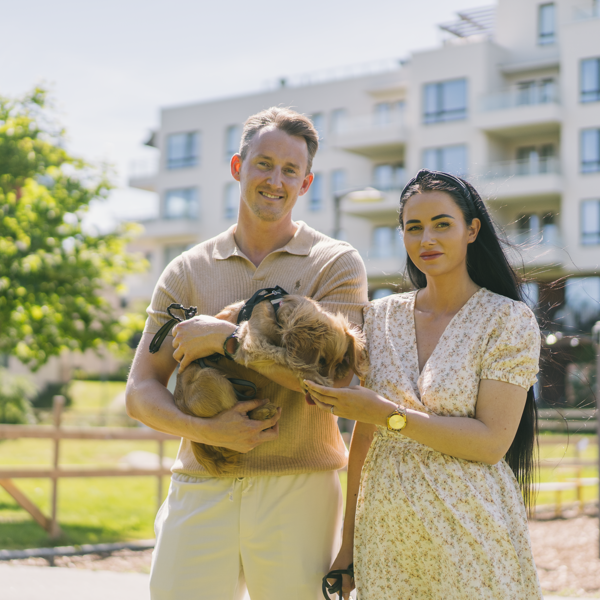 Dame og mann som står med en hund i hendene utenfor et boligområde i byen. Sol og sommer. 