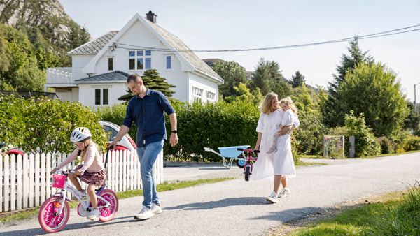 Mann og dame sammen med to små jenter som har med sykkel. Sommerlig antrukket. Hvitt eldre hus i bakgrunn. 