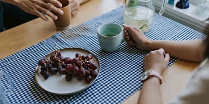 To personer sitter ved et kjøkkenbord, det er kun armene som er synlig. De holder hver sin kaffekopp og det står et fat med røde druer på bordet.