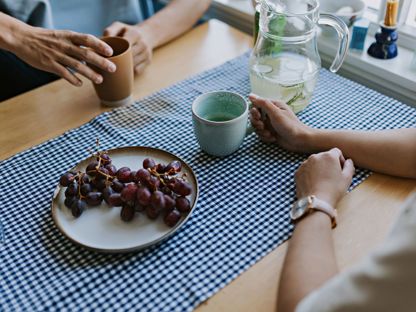 To personer sitter ved et kjøkkenbord, det er kun armene som er synlig. De holder hver sin kaffekopp og det står et fat med røde druer på bordet.