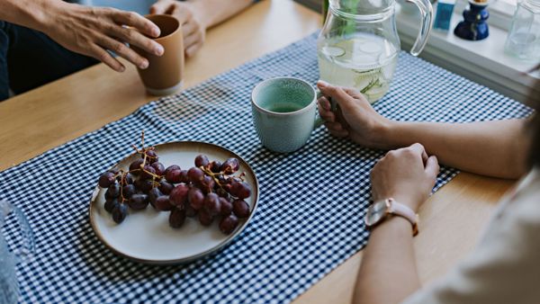To personer sitter ved et kjøkkenbord, det er kun armene som er synlig. De holder hver sin kaffekopp og det står et fat med røde druer på bordet.