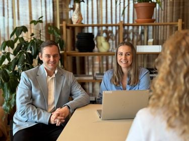 Kvinne og mann sitter bak et bord, ser inn i kamera og smiler. Kvinnen har en laptop fremfor seg. Det sitter en annen kvinne ved bordet, med ryggen mot kamera.