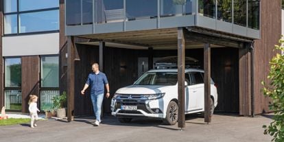 Mann og barn utenfor moderne hus, der bilen står parkert under veranda. Sol og vårlig kledde.