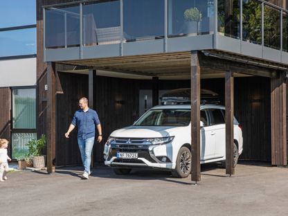 Mann og barn utenfor moderne hus, der bilen står parkert under veranda. Sol og vårlig kledde.