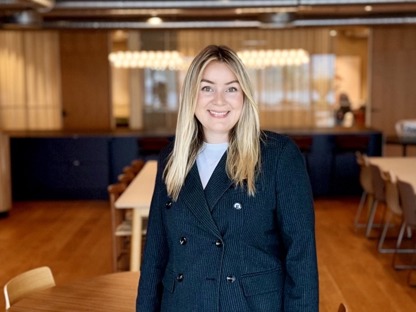 Dame med dressjakke som smiler til kamera, moderne lokale i bakgrunn. 