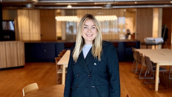 Dame med dressjakke som smiler til kamera, moderne lokale i bakgrunn. 