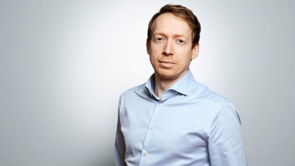 Portrett av Christian Balsnes i lyseblå skjorte mot en lys grå bakgrunn. Han har et rolig uttrykk og ser direkte mot kamera.