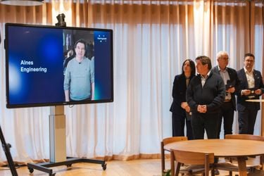 Stor tv-skjerm på stativ som viser tekst og bilde. Flere mennesker i høyre hjørnet på bildet ser mot denne skjermen. 