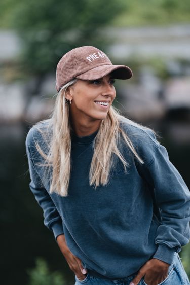 Kvinne med langt lyst hår og caps. Smiler og ser på noen utenfor bildet. Lener seg lett fremover og har hendene i lommen. 