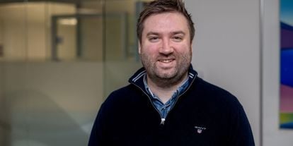 En smilende mann med skjegg og kort, brunt hår iført en mørk blå genser med en skjortekrage synlig under. Han står i et moderne kontormiljø med glassvegger i bakgrunnen.