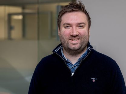En smilende mann med skjegg og kort, brunt hår iført en mørk blå genser med en skjortekrage synlig under. Han står i et moderne kontormiljø med glassvegger i bakgrunnen.