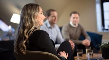 En smilende kvinne med langt, bølget hår sitter i en stol iført en mørk blazer. I bakgrunnen sitter to menn i uformelle klær, og de ser ut til å være i en avslappet samtale i et moderne og hyggelig kontormiljø.
