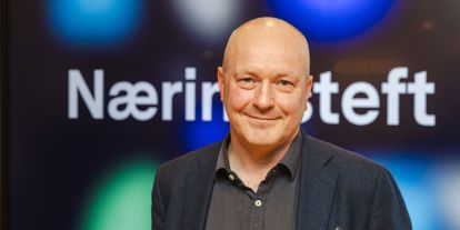 Mann uten hår, med dressjakke og skjorte ser i kamera. Skjerm med Næringsteft bak. 