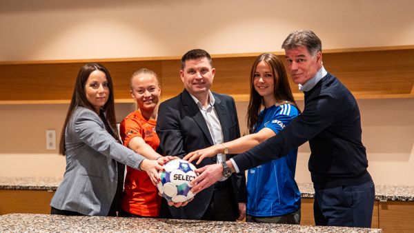 Three women, two with football shirts, and two men stand together and hold a hand each on a football.