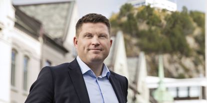 Dresskledd mann, stående med fjellstua i Ålesund bak seg. 