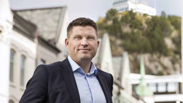 Dresskledd mann, stående med fjellstua i Ålesund bak seg. 