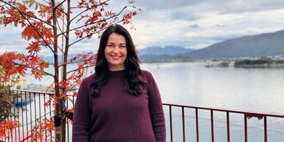 Dame med mørkt hår med fjell og fjord i bakgrunn. Ute på veranda, smiler i kamera