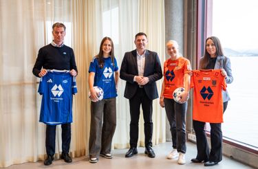 Five people stand in a row and smile at the camera. Two women are wearing football shirts and holding a ball. Two are holding suits in their hands.