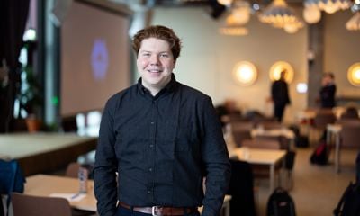 Ung mann med mørk skjorte, hendene i lommen. Smiler til kamera. Blurret bakgrunn