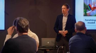Christian Balsnes holder en presentasjon foran en gruppe deltakere under et faglig seminar. Han står ved siden av en skjerm med en forretningsmodell og et bilde av fargepenner, og formidler tydelig i en profesjonell setting