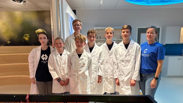 Skulebarn med hvite labfrakker står sammen med en dame i blå t-skjorte. De står foran et robotbord for legoroboter. 