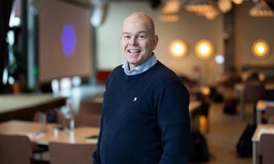 Mann med mørk blå genser og skjorte, smiler og ser i kamera. Blurret bakgrunn. 