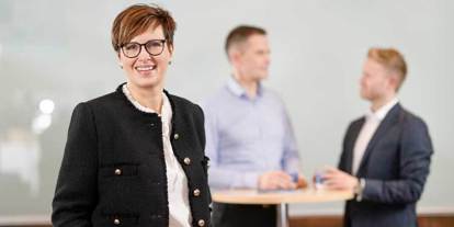 Smilende kvinne i sort jakke og briller står i fokus, mens to menn i dress og skjorte snakker ved et ståbord i bakgrunnen.
