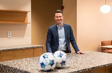 Mann med dress står ved en kjøkkenbenk med hendene på platen. I front av bildet ligger to fotballer. 
