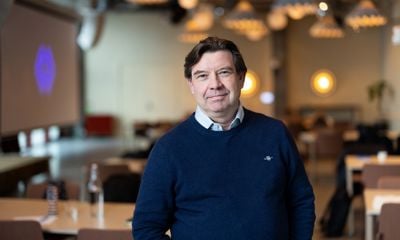 Mann med mørkt hår, mørk genser med skjorte under. Smiler og ser i kamera. Blurret bakgrunn