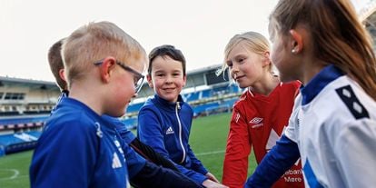 Gutter og jenter som står i en ring med hendene inn i midten, på en fotballbane. En gutt ser i kamera. 