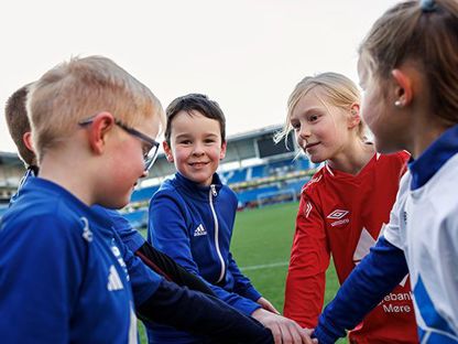 Gutter og jenter som står i en ring med hendene inn i midten, på en fotballbane. En gutt ser i kamera. 