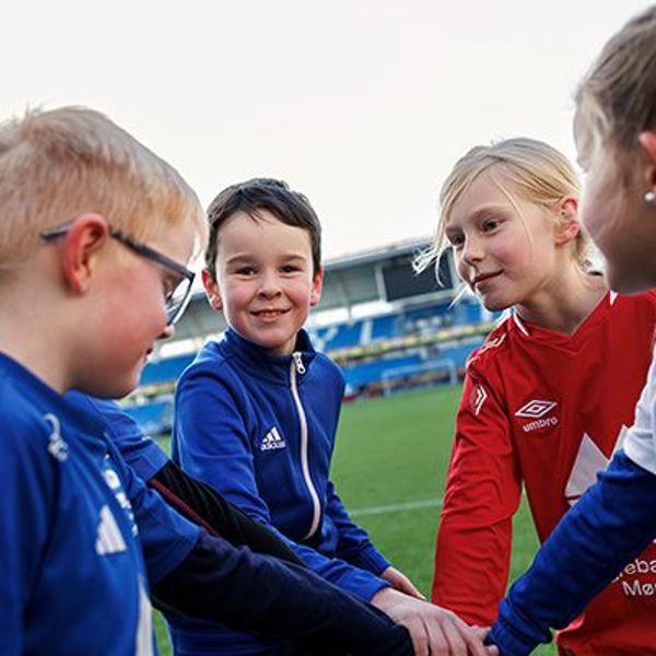 Gutter og jenter som står i en ring med hendene inn i midten, på en fotballbane. En gutt ser i kamera. 