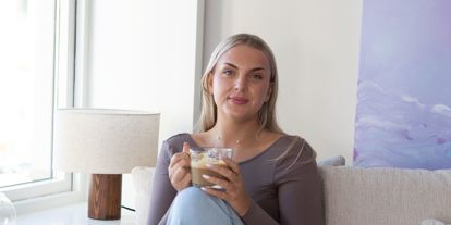 Ung kvinne i et moderne innemiljø, i sofa med en kopp kaffi. Smiler til kamera. 
