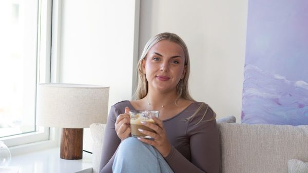 Ung kvinne i et moderne innemiljø, i sofa med en kopp kaffi. Smiler til kamera. 