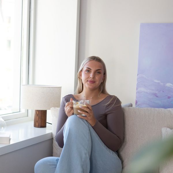 Ung kvinne i et moderne innemiljø, i sofa med en kopp kaffi. Smiler til kamera. 