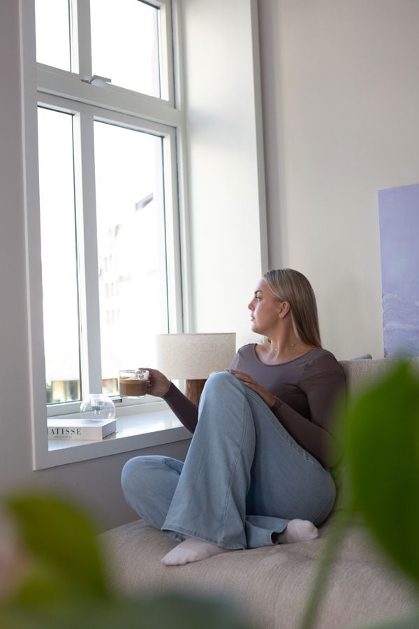 Ung kvinne som er ut av vinduet med kaffi i hånden, moderne leilighet i by. 