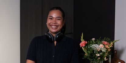 Ung kvinne med headset rundt halsen, på kjøkkenet. Hun står oppreist og smiler til kamera