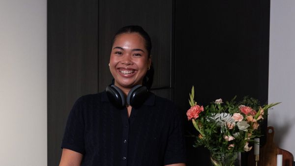 Ung kvinne med headset rundt halsen, på kjøkkenet. Hun står oppreist og smiler til kamera