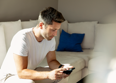Mann i hvit T-skjorte sitter på en sofa og bruker mobiltelefon, med hvite puter og en blå pyntepute i bakgrunnen.